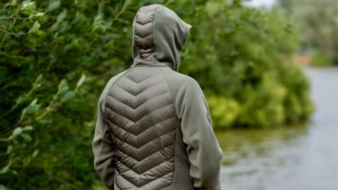 Person viewed from behind wearing a quilted olive-green jacket with a chevron pattern, standing near a body of water and lush greenery