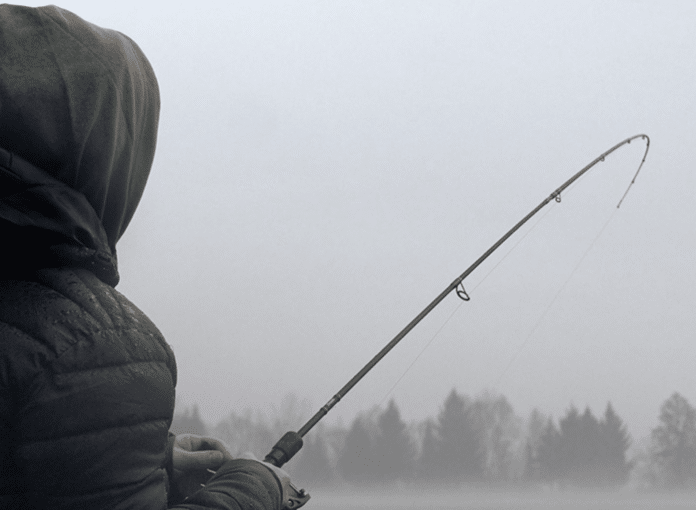 Person in a green hooded jacket holding a fishing rod beside a misty lake in winter.