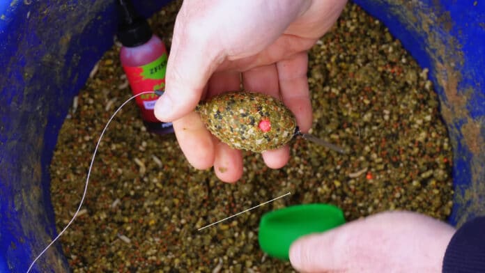Person holding a dense groundbait ball with a visible hook embedded, in a blue tub with fishing attractant bottle nearby.