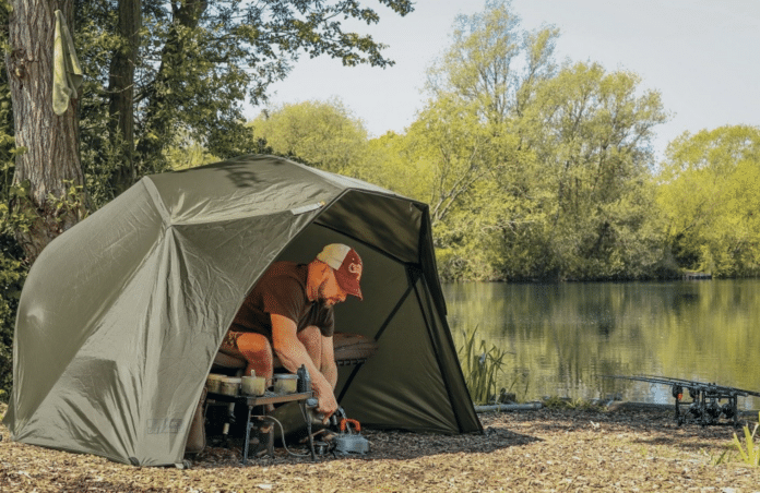 Man camping by a lake, cooking gear under a green tent near trees by the water.