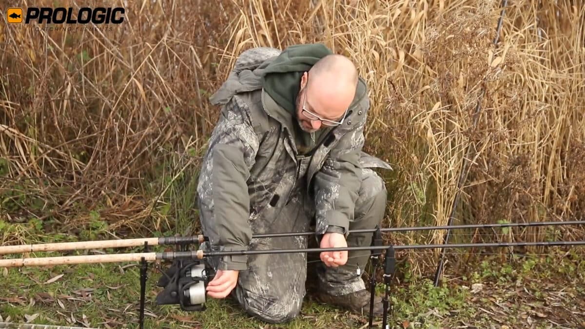 Man in camouflage kneeling outdoors among tall reeds, handling fishing rods and reel. (Prologic logo appears in the corner)