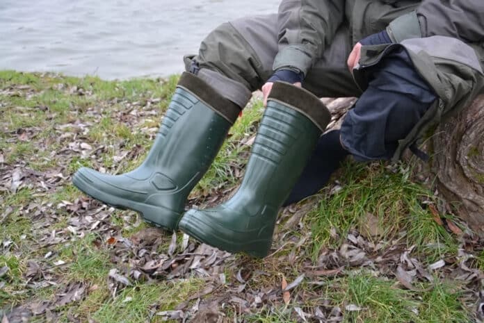 Person in green rain boots sitting on a log by a lake, with grass and fallen leaves around.