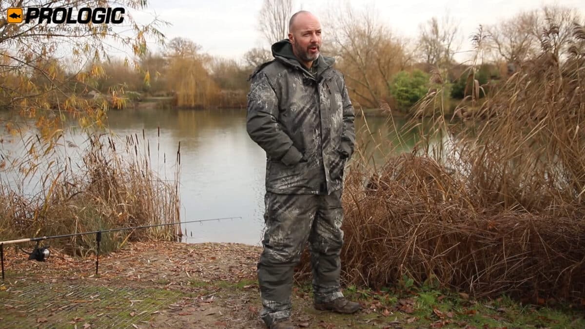 Man in camouflage fishing jacket and pants stands by a lake among autumn reeds; Prologic logo in the top-left corner.