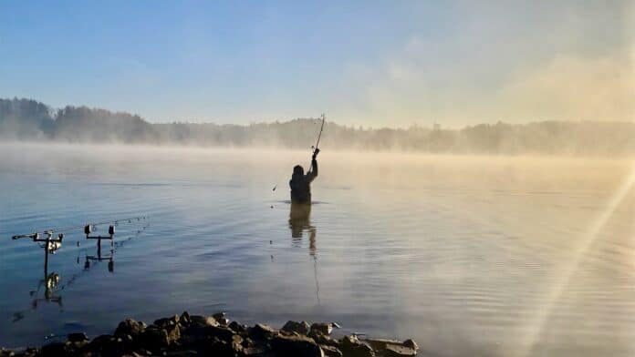 Person waist-deep in calm lake, raising a fishing rod as mist rises from the water at dawn