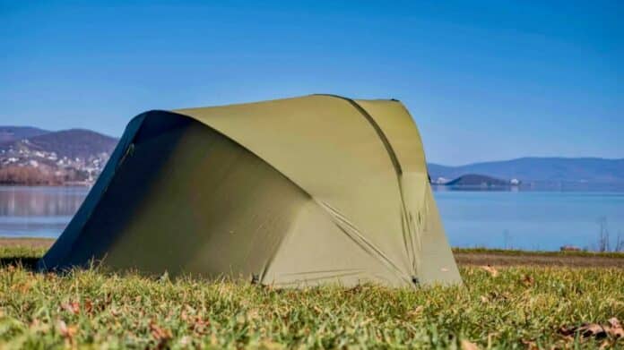 Green tent pitched on grassy lakeside with blue water and distant hills under a clear sky.