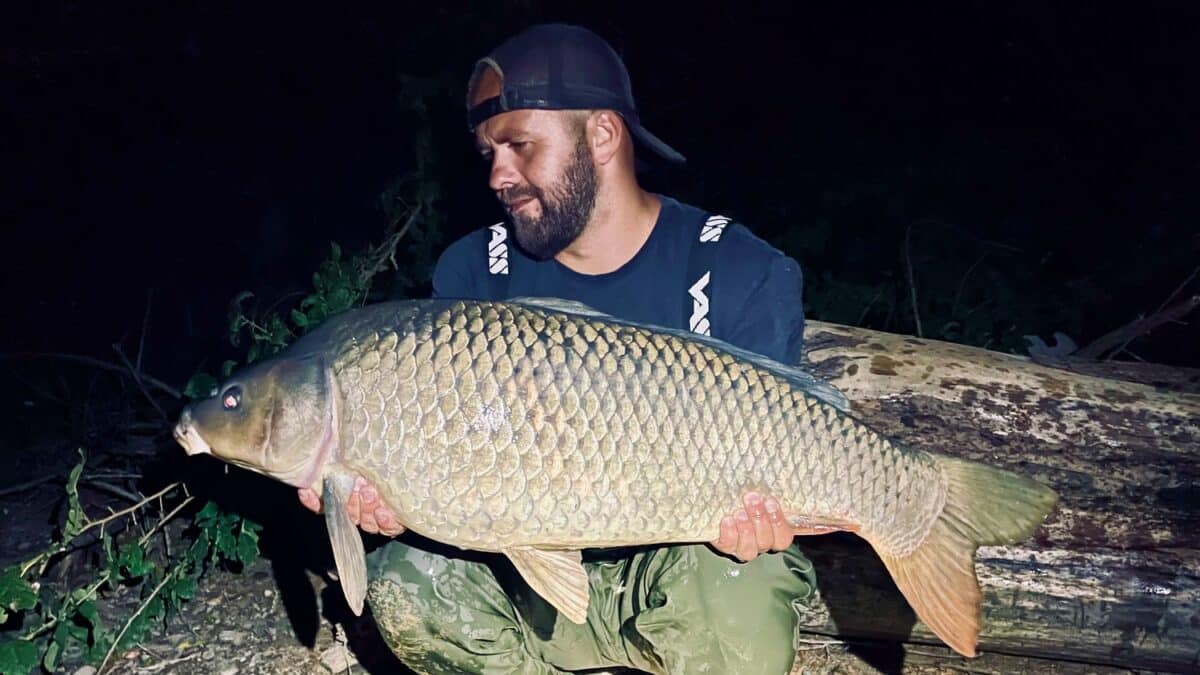 Man at night wearing a backward cap and dark shirt holding a large carp beside a fallen log, posing for a photo.