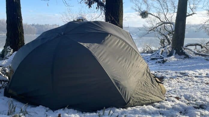 Dark green dome tent pitched on snow beside a calm lake, surrounded by leafless trees in a winter campsite.