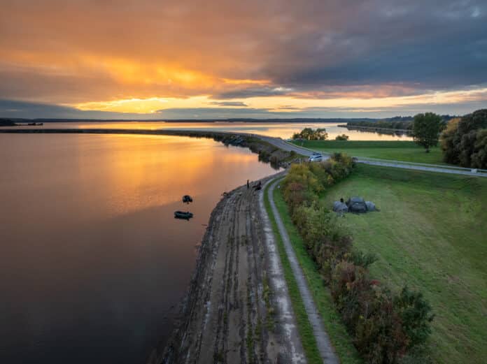 Aerial view of a river at sunset with orange sky, a stepped rocky embankment along the water, and a green park with tents on the right edge