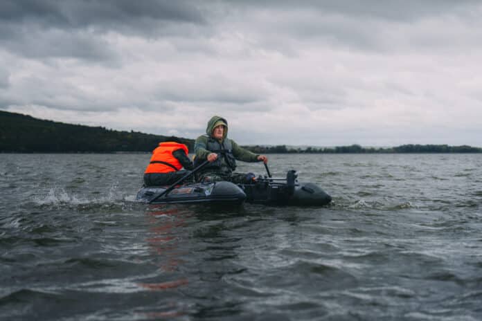 Two people in a small inflatable boat on choppy lake water under a gray, cloudy sky; one wears a bright orange life jacket while steering.