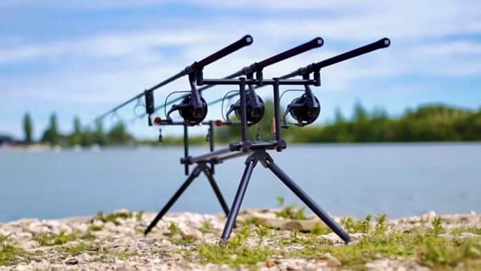 Three fishing rods secured in a tripod rod holder by a lakeside.