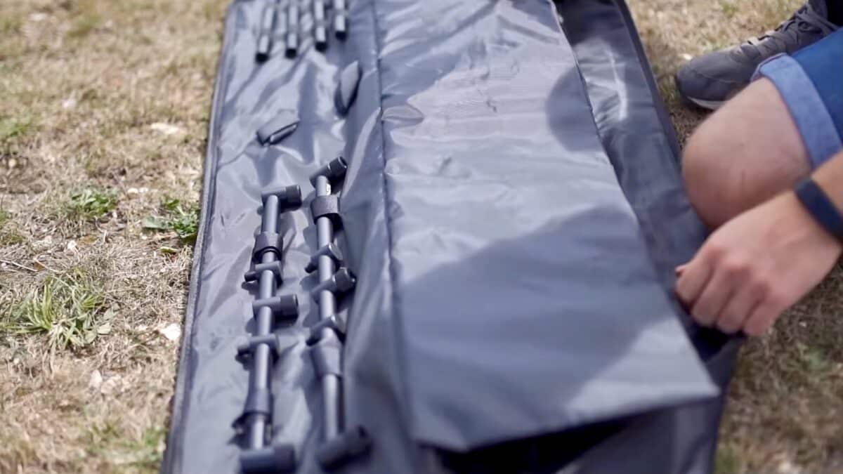Outdoor scene with a grey gear bag laid open on grass and foldable trekking poles clipped inside, a person’s leg at right edge.]