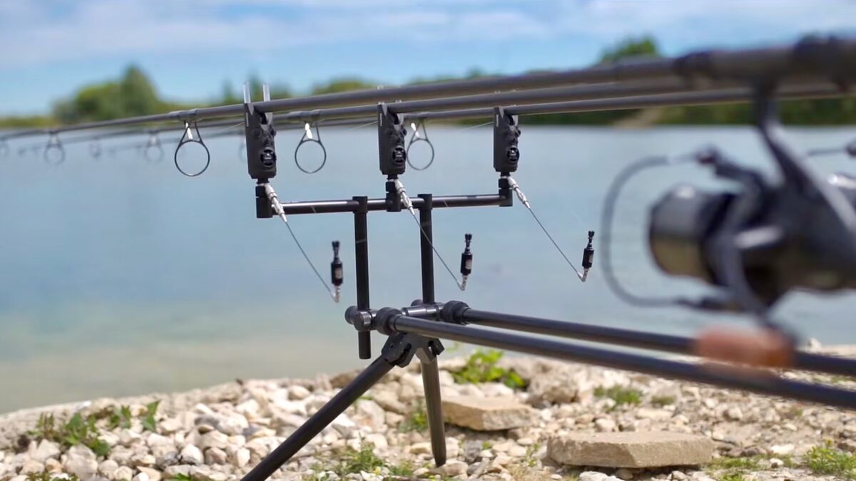 Fishing rod rig with multiple reels on a tripod by a rocky lakeshore.