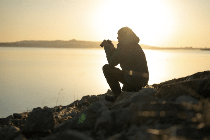Person in a hooded jacket crouches on a rocky shoreline, binoculars raised to survey the sunset-lit water.