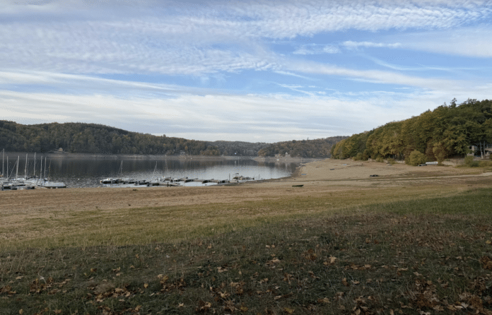 Calm lake with sailboats docked along a small marina and tree-covered hills in the background on a clear day at autumn shore.