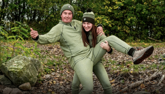 Two people in matching olive-green sweaters and beanies pose playfully outdoors, with one person lifting the other in a forest setting.