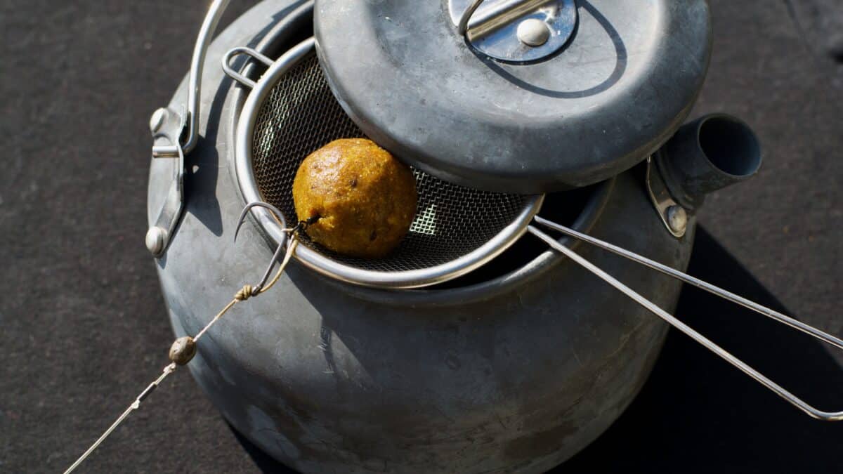 Close-up of a gray metal container with a circular mesh insert; a small brown, textured ball sits inside, tied with string.