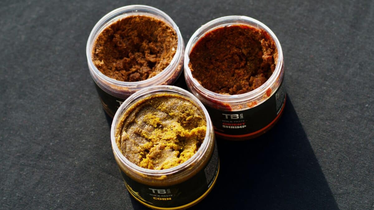 Four open jars of boilie paste in brown, red, and yellow hues on a dark surface, viewed from above.