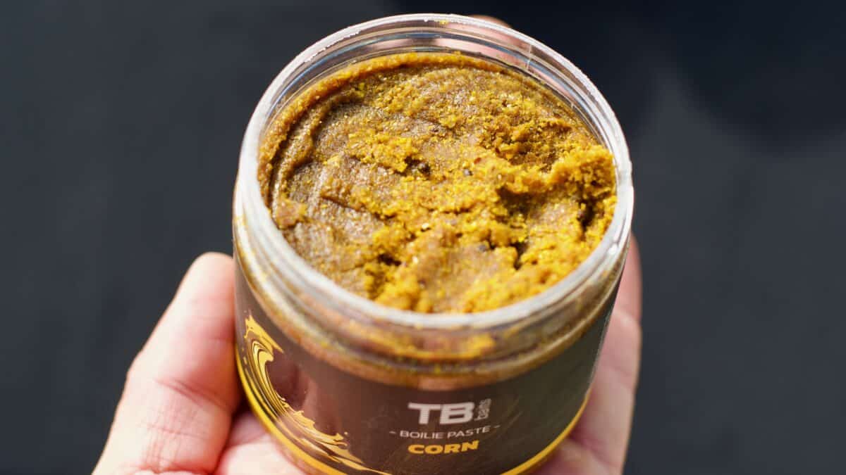 Close-up of a hand holding an open jar of brown corn paste, label showing the word 'CORN' on a dark sleeve.