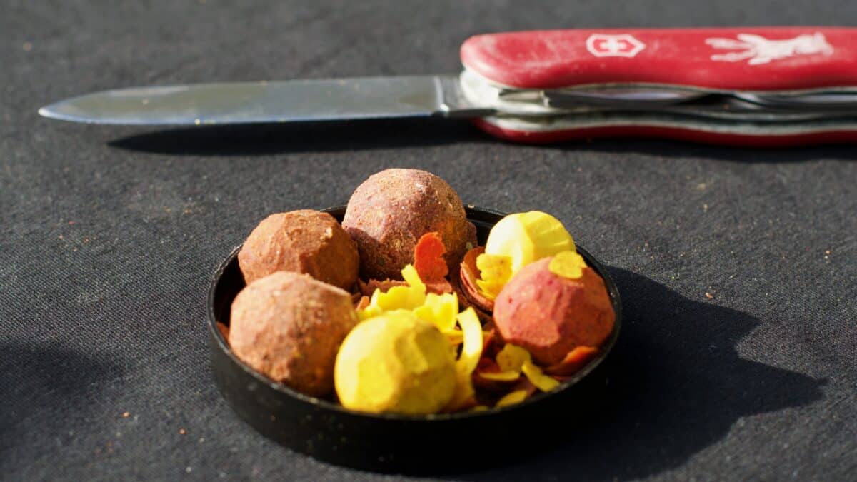 Red-handled pocket knife with blade extended sits above a bowl of colorful meatballs and yellow zest on a dark tablecloth to the left of the dish.