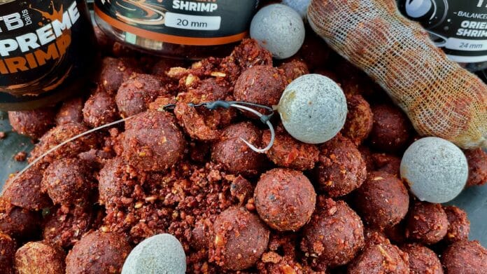 Pile of red fishing bait boilies with bits of seasoning, gray fishing weights, and a fishing net bag in the background.