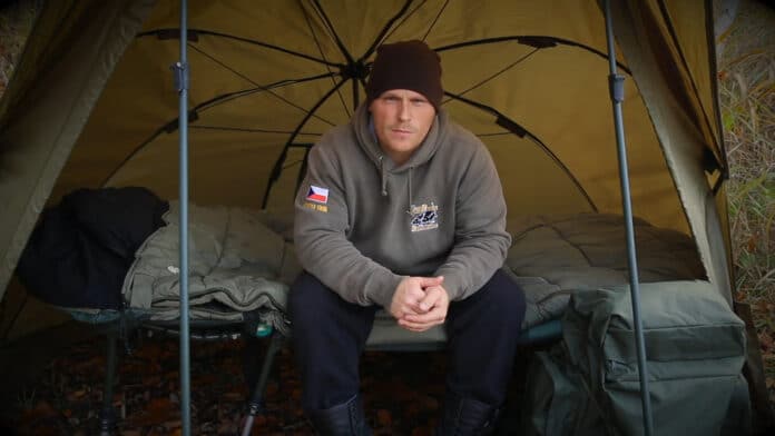 Man in a gray hoodie and black beanie sits on a camping cot inside a yellow tent, hands clasped and looking at the camera.