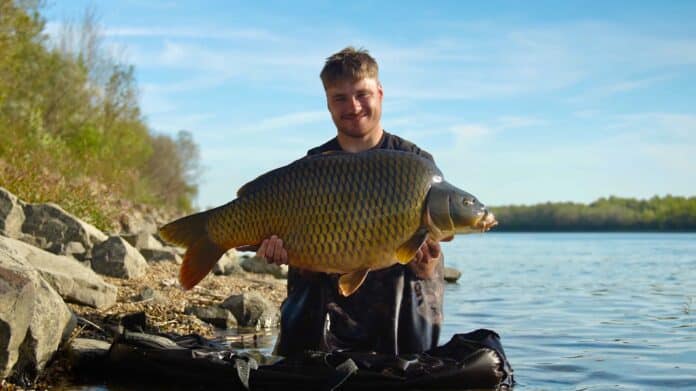 Smiling man holds a large golden fish across his chest on a rocky lakeshore near calm water and trees.