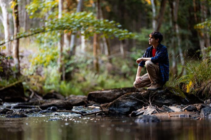 Young man crouching on a rock by a forest stream, looking into the distance.